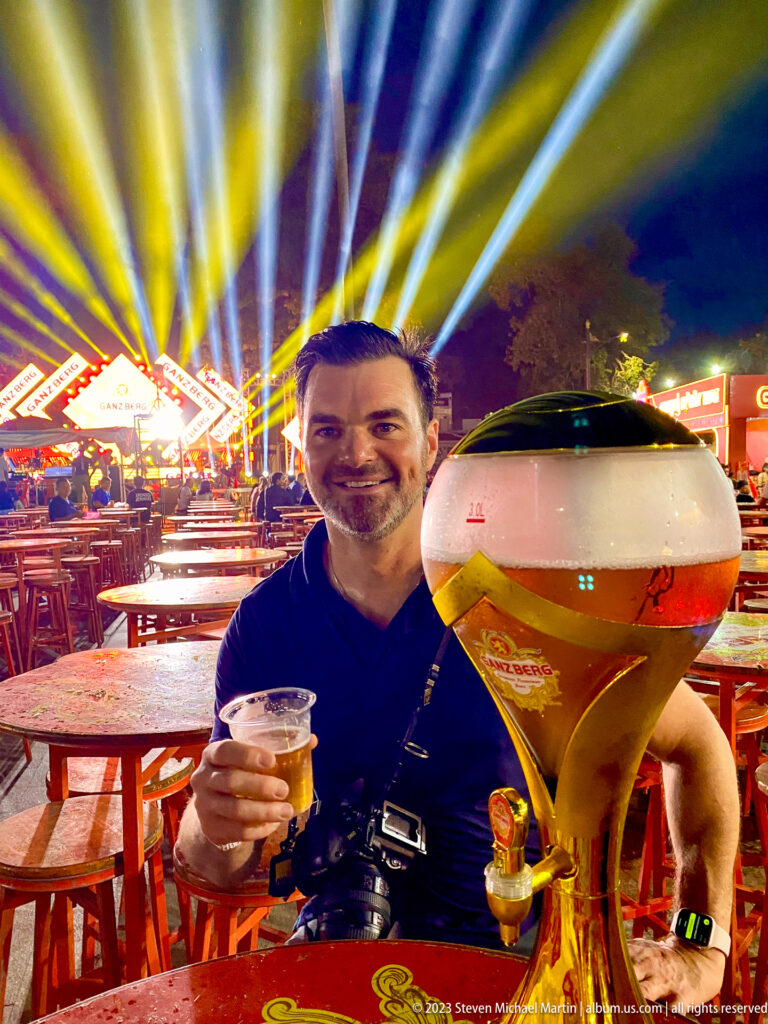 Smiling man holding a beer beside a large dispenser with stage lights and market tables behind.