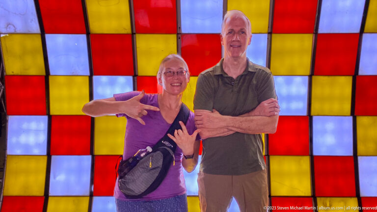 Two smiling adults posing with crossed arms before red, yellow, and white illuminated panels.