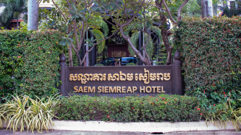 Wooden Saem Siemreap Hotel sign in Khmer and English surrounded by dense shrubs and trees.