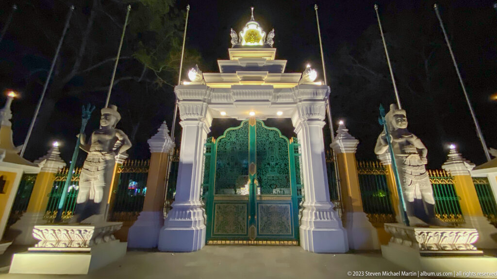 Wide nighttime view of white gateway, green gates, and two guardian statues holding staffs.