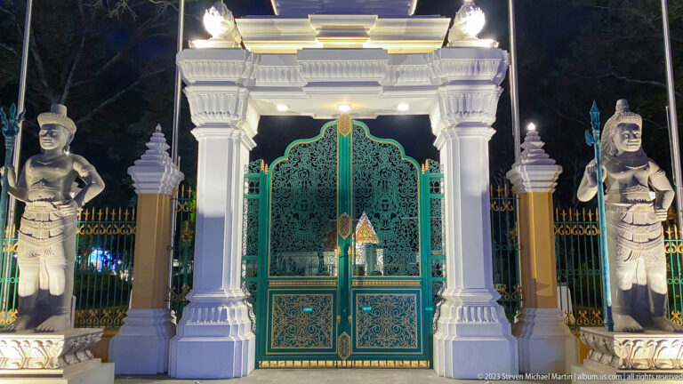 Frontal nighttime view of ornate green gates beneath a white gateway, flanked by guardian statues.