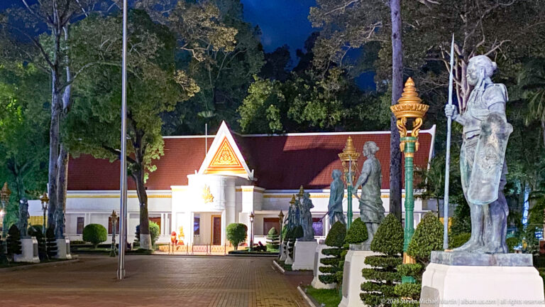 Nighttime avenue lined with statues and ornate lamps leading to a brightly lit gabled building.