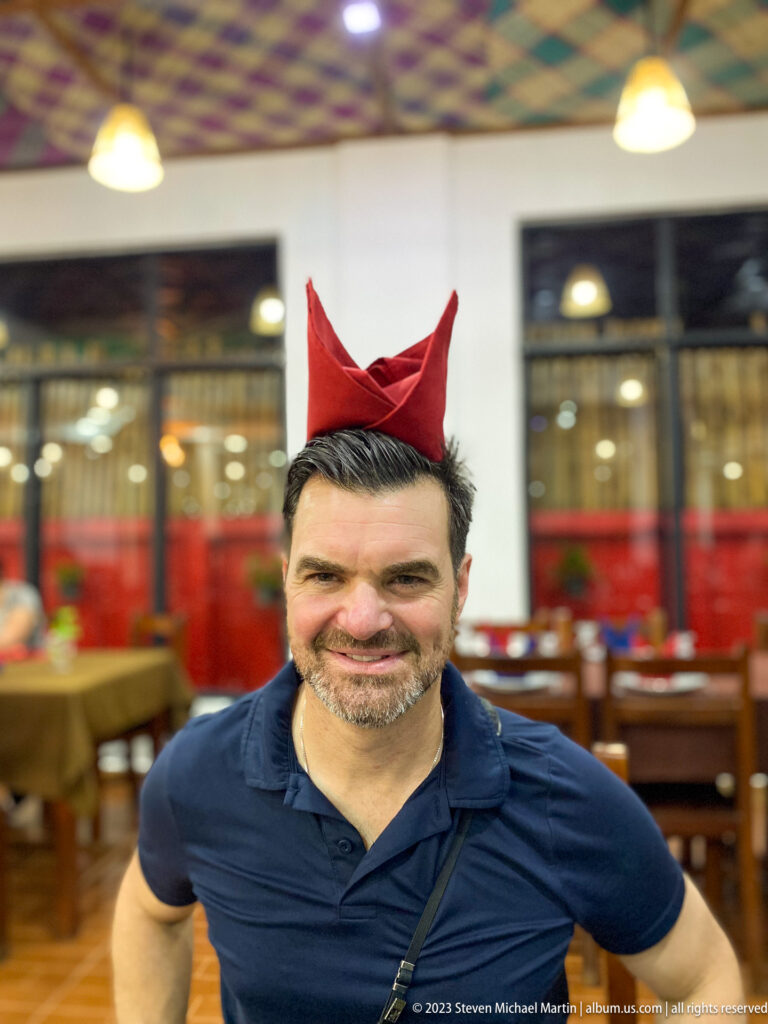 Smiling man in a restaurant wearing a folded red napkin as a hat.