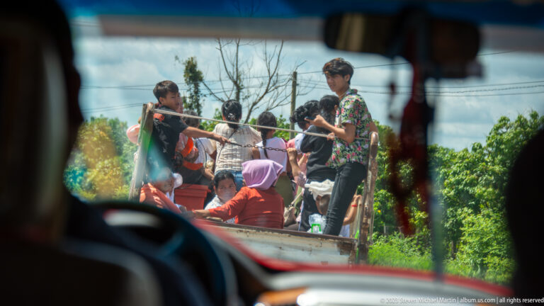 Passengers standing and sitting in an open vehicle, seen through a windshield on a rural road.