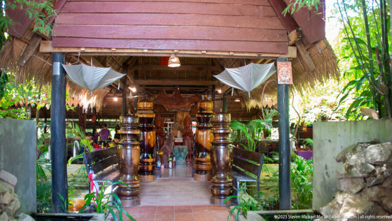 Thatched restaurant entrance with polished wooden columns, benches, and dense tropical plants.