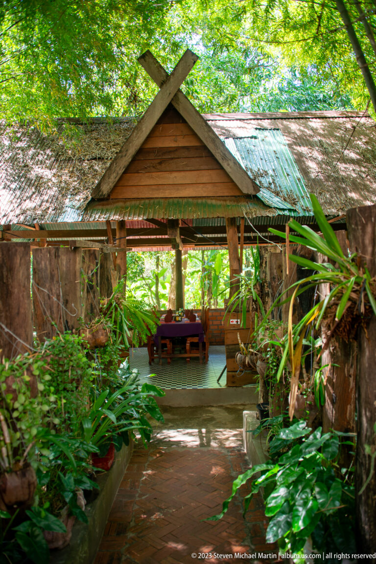 Small open-sided wooden pavilion with gabled roof, bench, and dense tropical foliage.