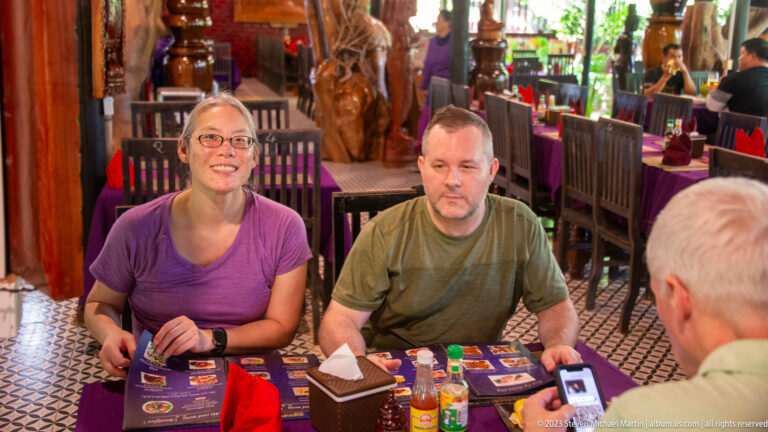 Two visitors seated at a restaurant table with menus and condiments, while a third person faces them.