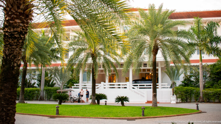 Long white hotel building with verandas and red roof behind palm trees and manicured lawn.