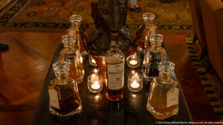 Macallan 18 whisky bottle surrounded by crystal decanters, glasses, candles, and a dark sculpture.