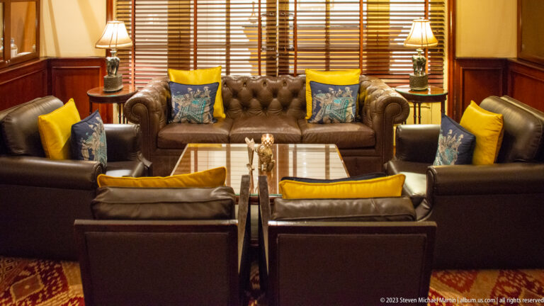 Dark leather sofas and armchairs with yellow and blue cushions around a glass table in a wood-paneled lounge.