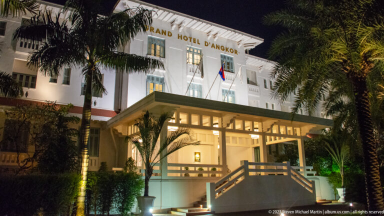 Illuminated white Grand Hotel d’Angkor entrance with covered stairs, palms, and Cambodian flag at night.