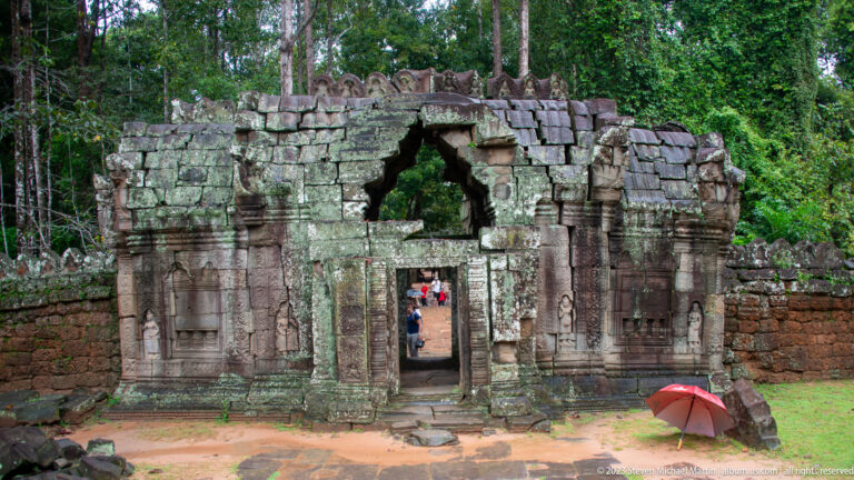 Ruined doorway with a collapsed triangular opening, carved niches, visitors, and a red umbrella.
