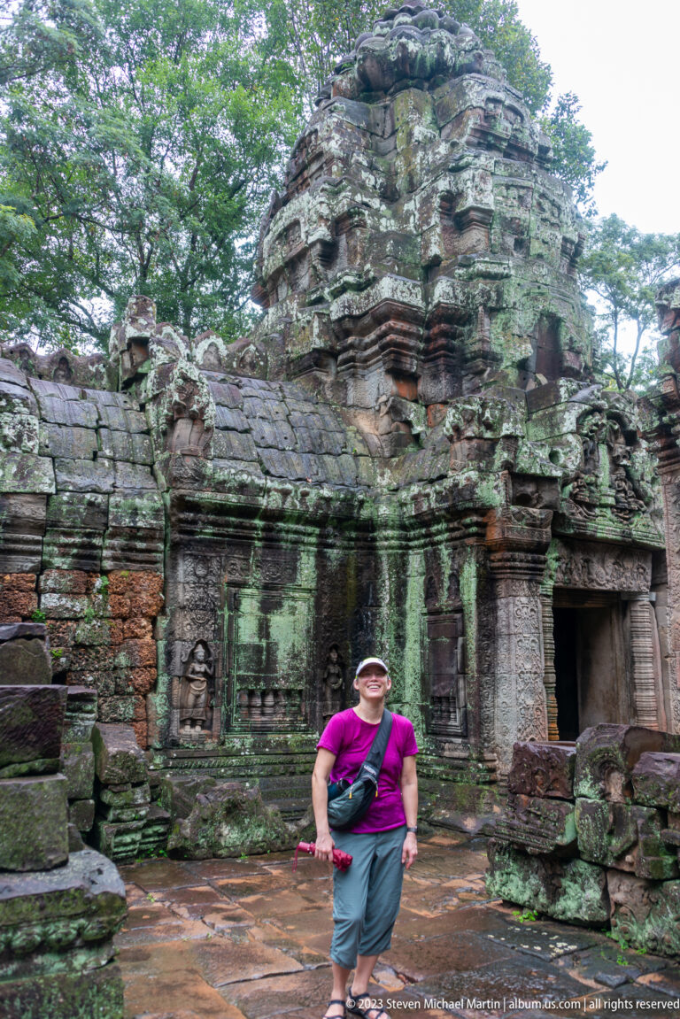 Visitor in a magenta shirt standing before weathered Ta Som masonry and low temple doorways.