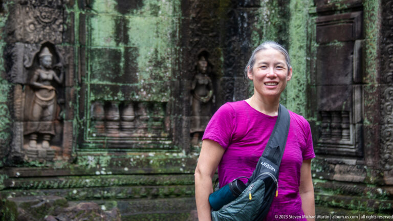 Smiling visitor in a magenta shirt before a dark doorway with carved female figures.