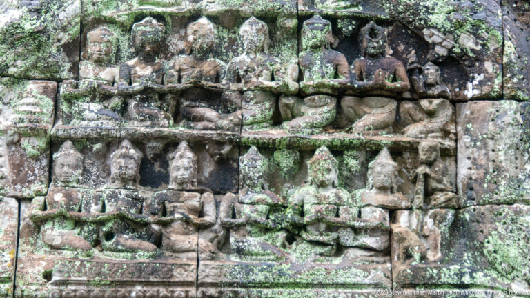Close view of a weathered sandstone relief with many small figures and heavy lichen.