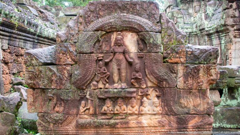 Standing carved figure beneath an ornate arch, with smaller figures along the base of the stone block.