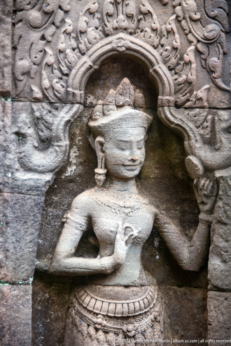 Carved female deity in an arched niche with one arm raised and decorative scrollwork beside her.
