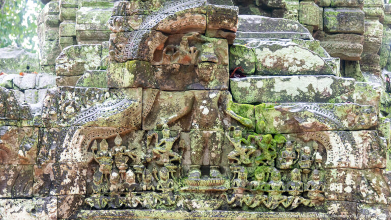 Weathered carved pediment above a doorway, with a central seated figure, attendants, and vegetal ornament.