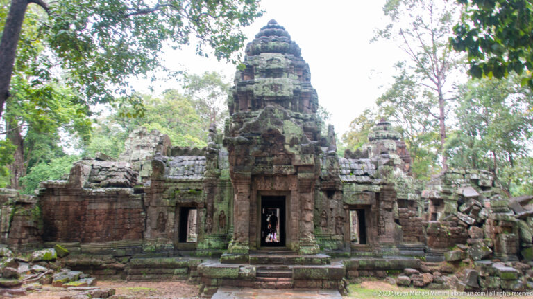 Frontal view of a Ta Som sanctuary with central tower, doorway, low side chambers, and surrounding trees.
