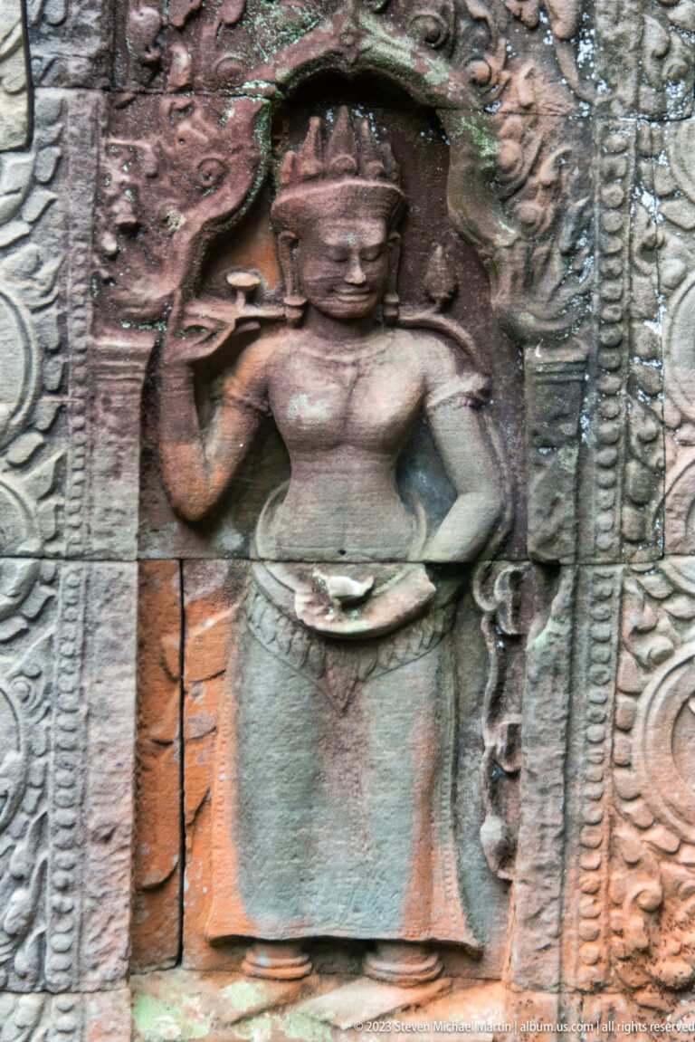 Standing female deity relief with raised hand, headdress, jewelry, and curling border ornament.
