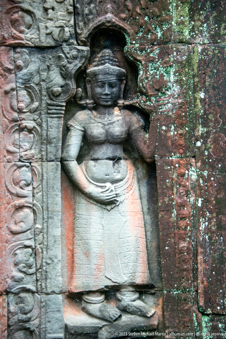 Weathered female deity relief with raised arm, ornate headdress, jewelry, and long garment.