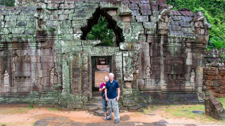 Two visitors standing in front of a ruined Ta Som doorway with a collapsed upper opening.