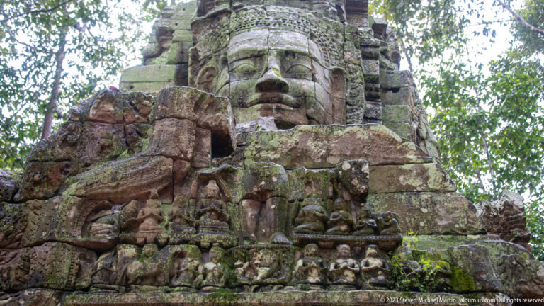 Close oblique view of a large carved face above a weathered pediment with multiple figures.