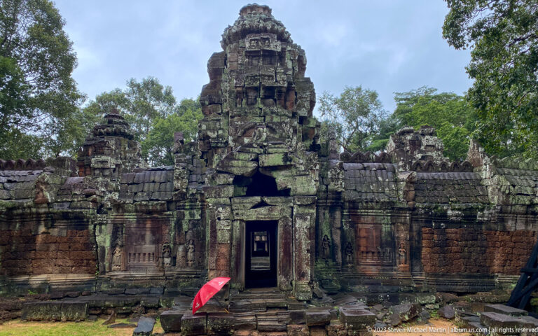 Frontal view of a towered Ta Som gateway with carved walls, dark doorway, and red umbrella.