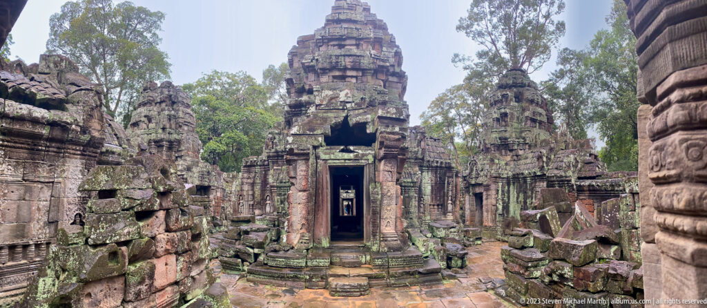 Wide oblique view of clustered Ta Som sanctuary towers, central doorway, broken walls, and scattered stones.