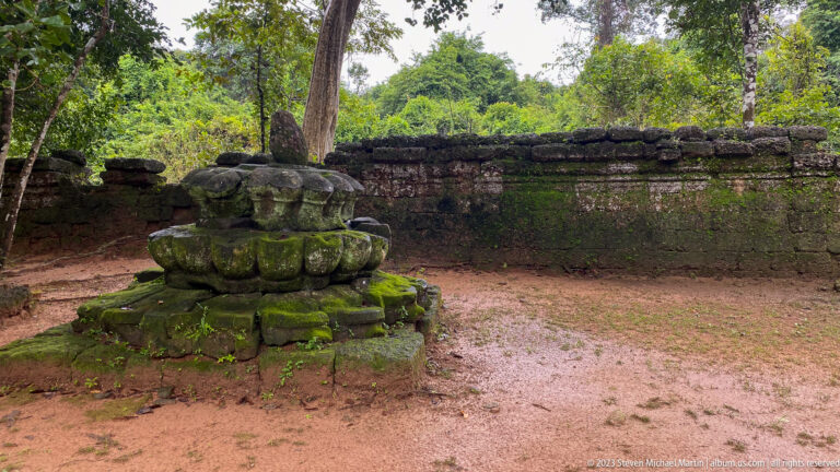 Moss-covered lotus-shaped stone base on bare ground beside a low temple enclosure wall.