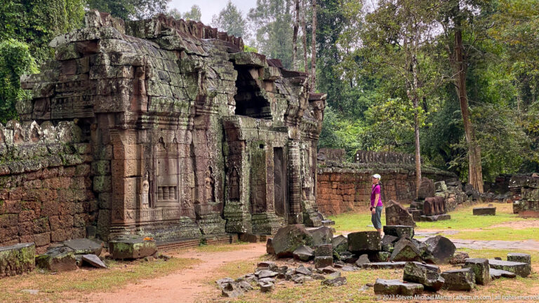 Visitor walking beside a small ruined gateway, scattered stones, and a long enclosure wall at Ta Som.