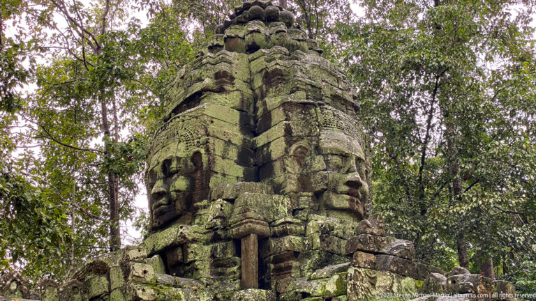 Low-angle corner view of a moss-covered Ta Som tower with multiple large carved faces.
