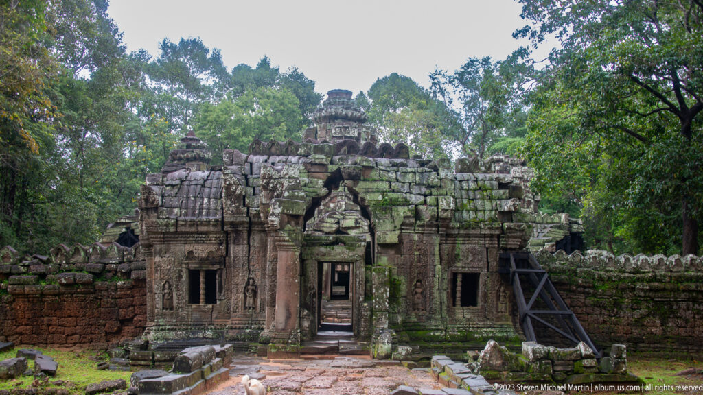 Wide view of a ruined Ta Som gateway with broken roofline, central doorway, and forest behind.