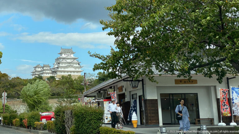 Himeji Castle’s white main keep and smaller towers rising above trees behind a low visitor building, hedges, signs, and pedestrians.
