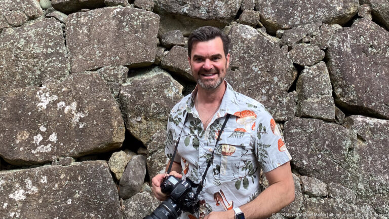Smiling traveler holding a camera in front of a massive wall of irregular gray stones at Himeji Castle.