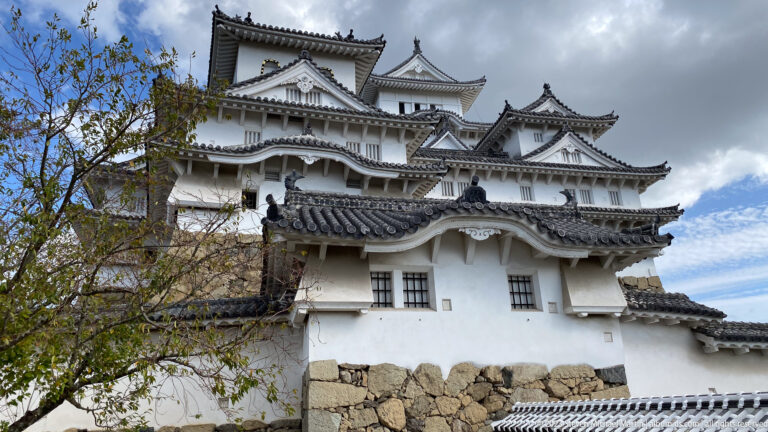 Upward view of Himeji Castle’s white-plastered turrets, layered gray tiled roofs, ornate gables, and stone base beneath a partly cloudy sky.