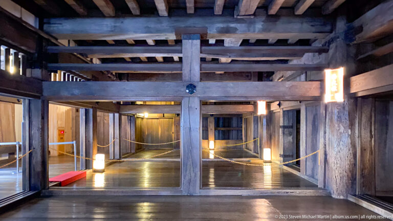 Interior of Himeji Castle showing massive wooden posts, horizontal beams, ceiling joists, polished floors, and roped visitor paths.