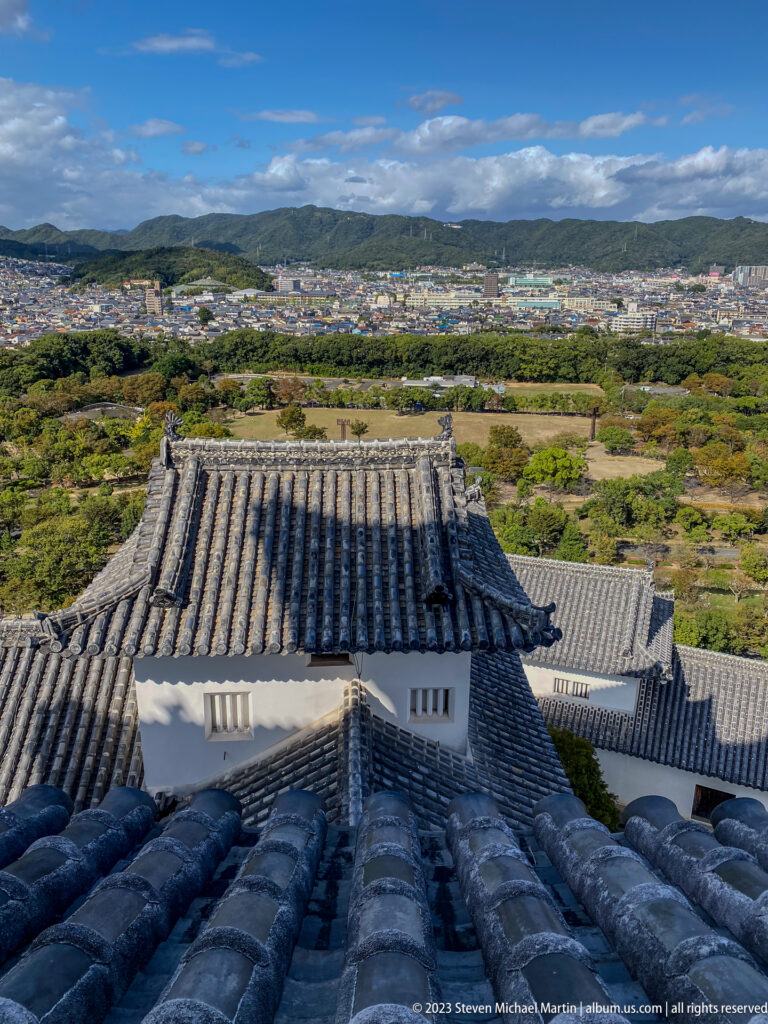Elevated view over layered gray tile roofs and white walls of Himeji Castle toward the city, fields, and low mountains under a blue sky.