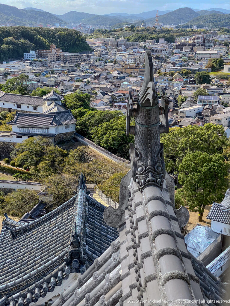 Dark sculptural roof ornament mounted on a gray tiled ridge, with Himeji’s cityscape and hills visible below.