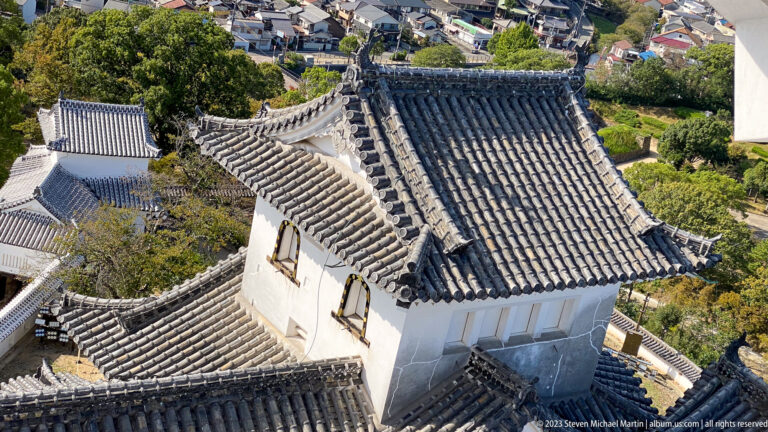 High-angle view of a white-plastered Himeji Castle building with a broad gray tiled roof, surrounded by lower roofs, walls, trees, and nearby houses.