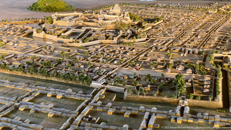 Large scale model of Himeji Castle surrounded by moats, walls, roads, buildings, and the dense street grid of the historic castle town.