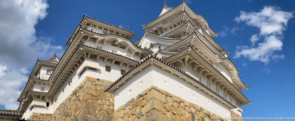 Low-angle view of Himeji Castle’s white main keep with layered gray roofs and ornate gables rising above a massive stone foundation under a blue sky.