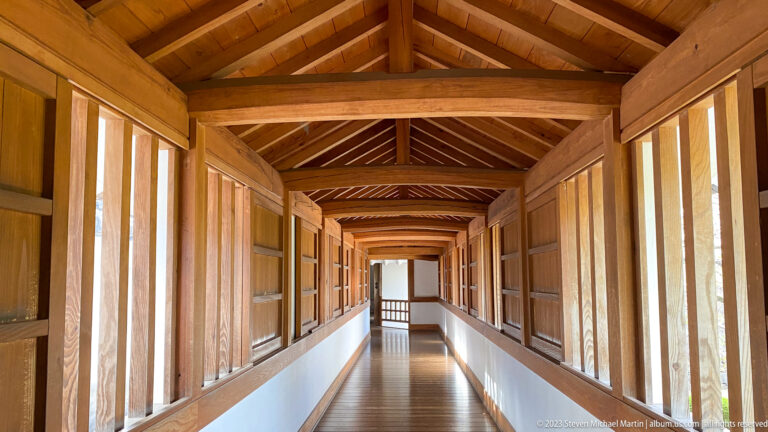 Long sunlit wooden corridor inside Himeji Castle with exposed rafters, repeating posts and windows, white lower walls, and a polished plank floor.