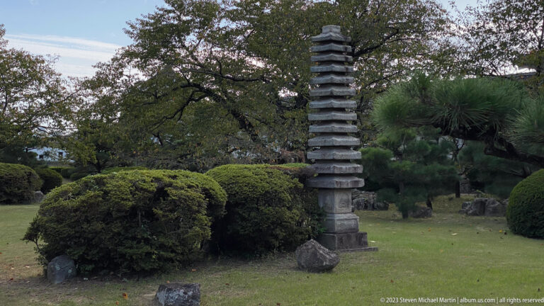 Tall multi-tiered stone pagoda standing on a lawn among clipped shrubs, pines, rocks, and mature trees near Himeji Castle.