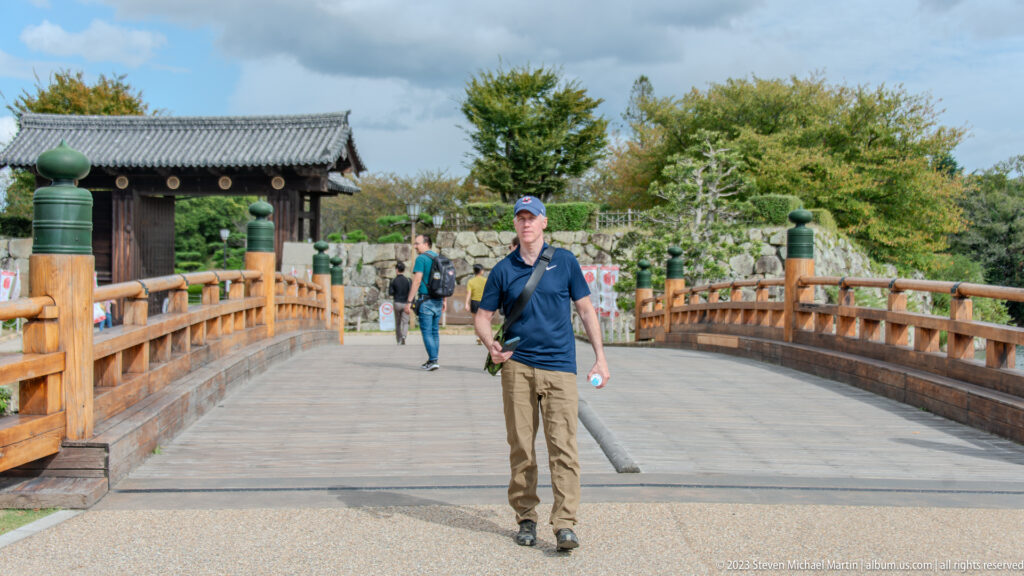 Visitors crossing a wooden bridge with green-capped posts toward a tiled gate at Himeji Castle.