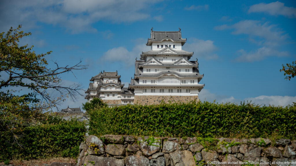 White main keep and smaller tower of Himeji Castle rising above hedges and a low stone wall.