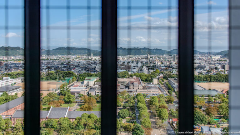 Himeji city panorama seen through dark vertical window bars from an upper castle floor.