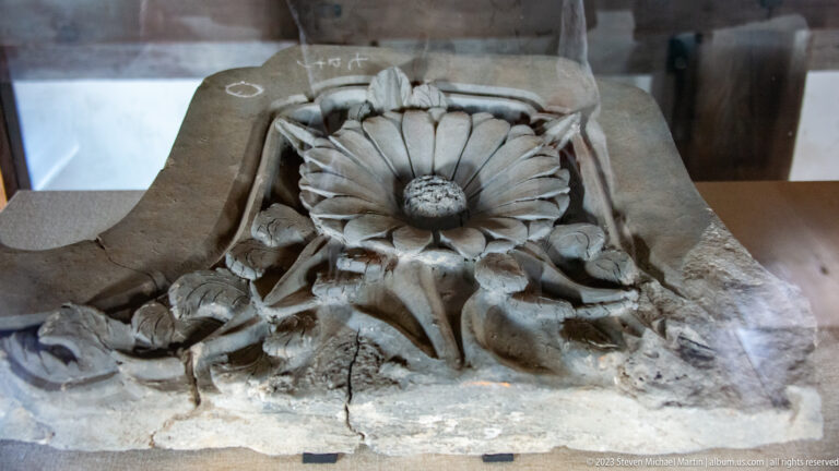Weathered floral architectural carving displayed in a museum case.