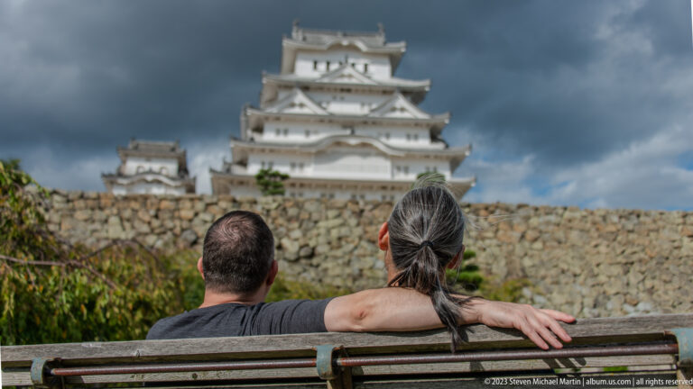 Two visitors seated on a bench facing Himeji Castle’s main keep beyond a stone wall.