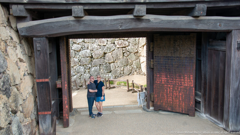 Two visitors beneath a heavy wooden gate set between stone walls at Himeji Castle.